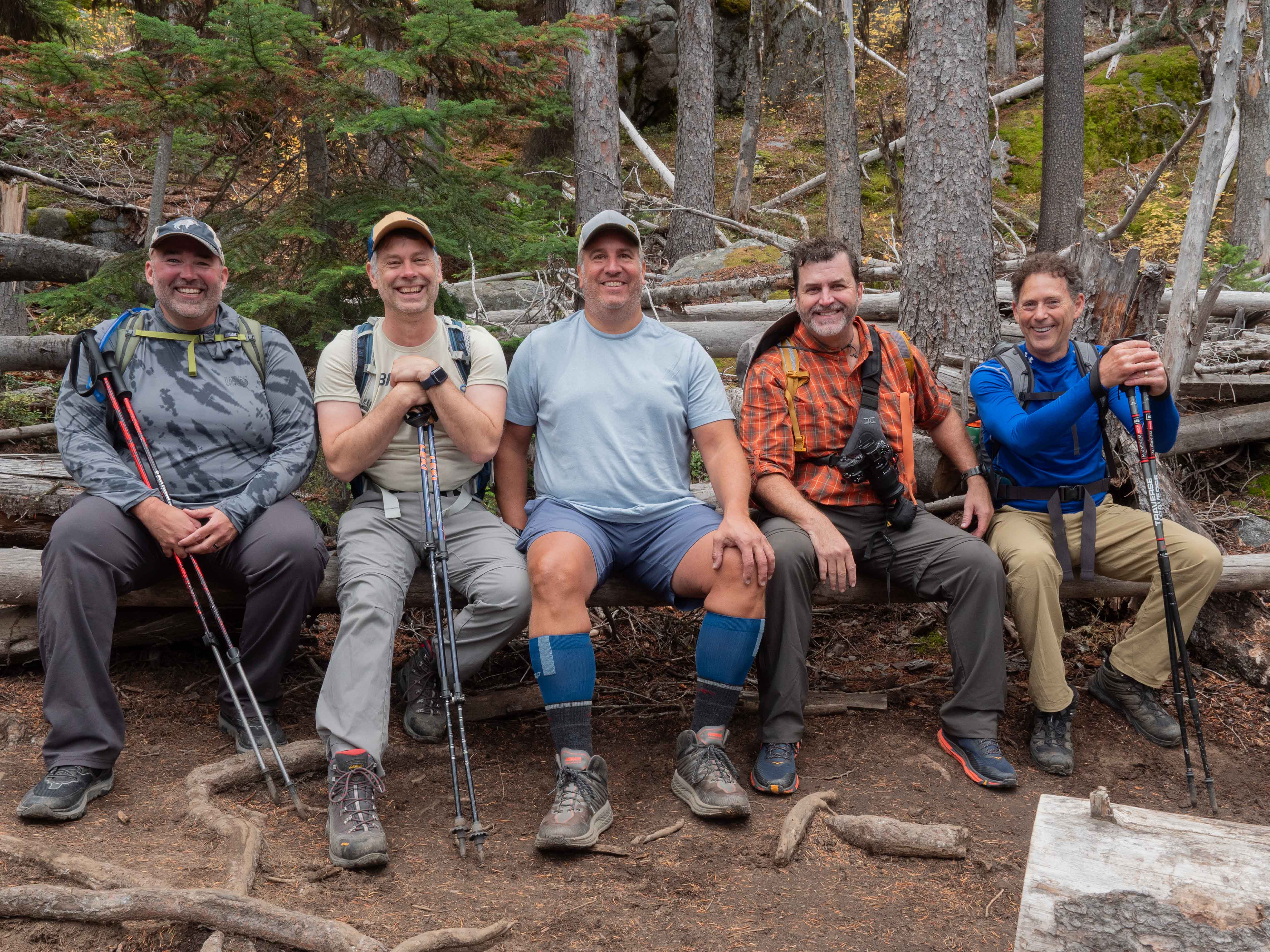 Group of five hikers resting on a log bench along the forested Colchuck Lake approach trail