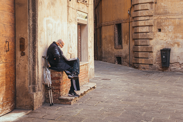 An elderly man reads a newspaper on a narrow medieval alley in Siena, warm stone walls behind him