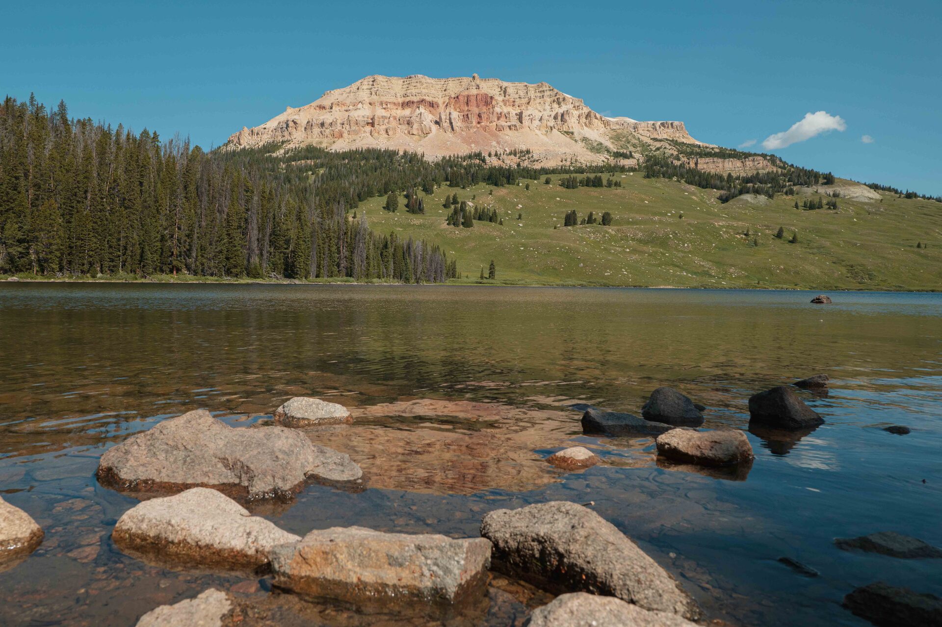 Beartooth Lake with rounded boulders in the crystal-clear shallows, a sweeping green hillside and the dramatic red and tan layered cliffs of Beartooth Butte rising above the far shore under a blue summer sky